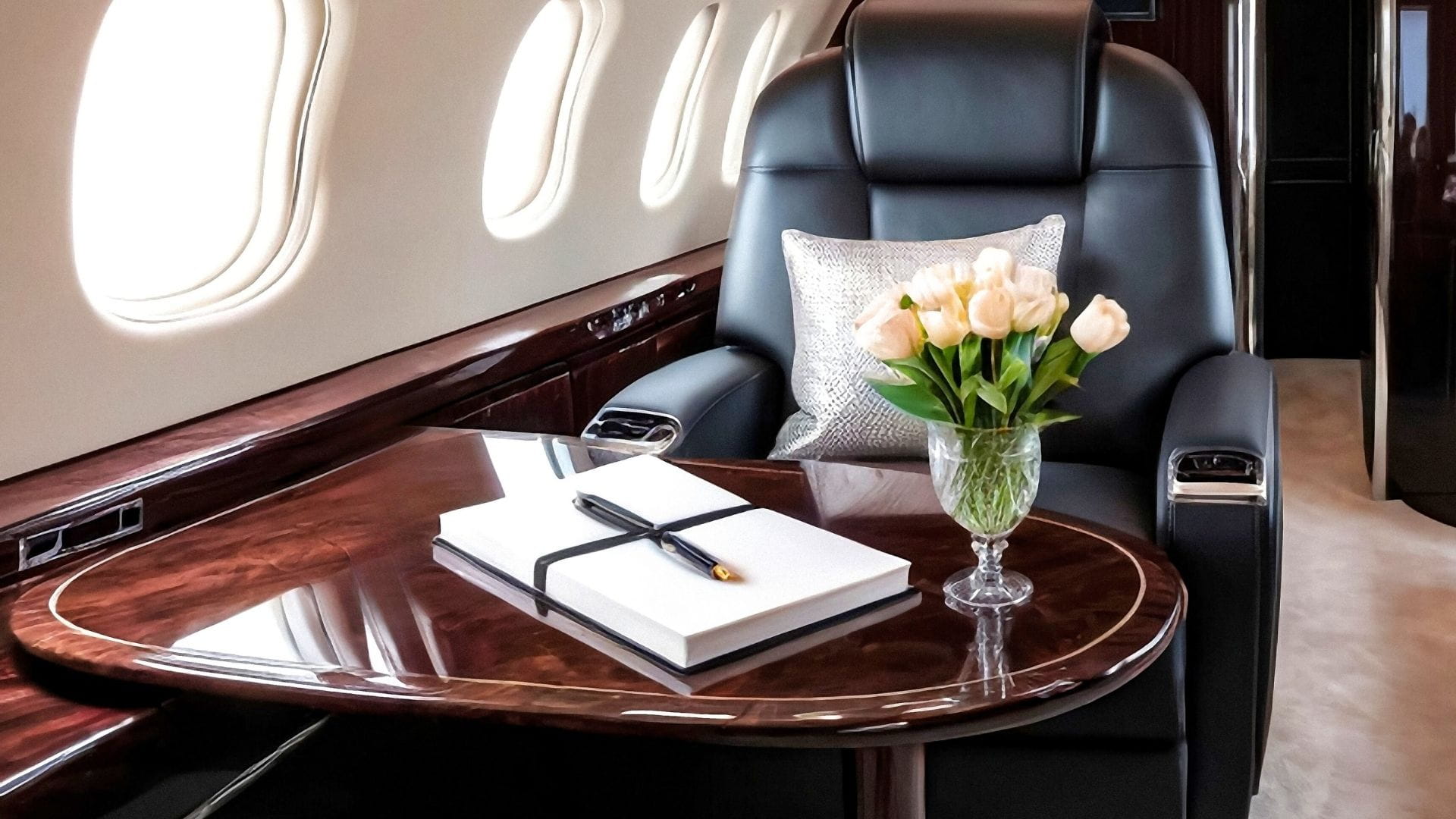 Private jet cabin interior with black leather seat, polished wood table, white guestbook and crystal vase of white tulips.